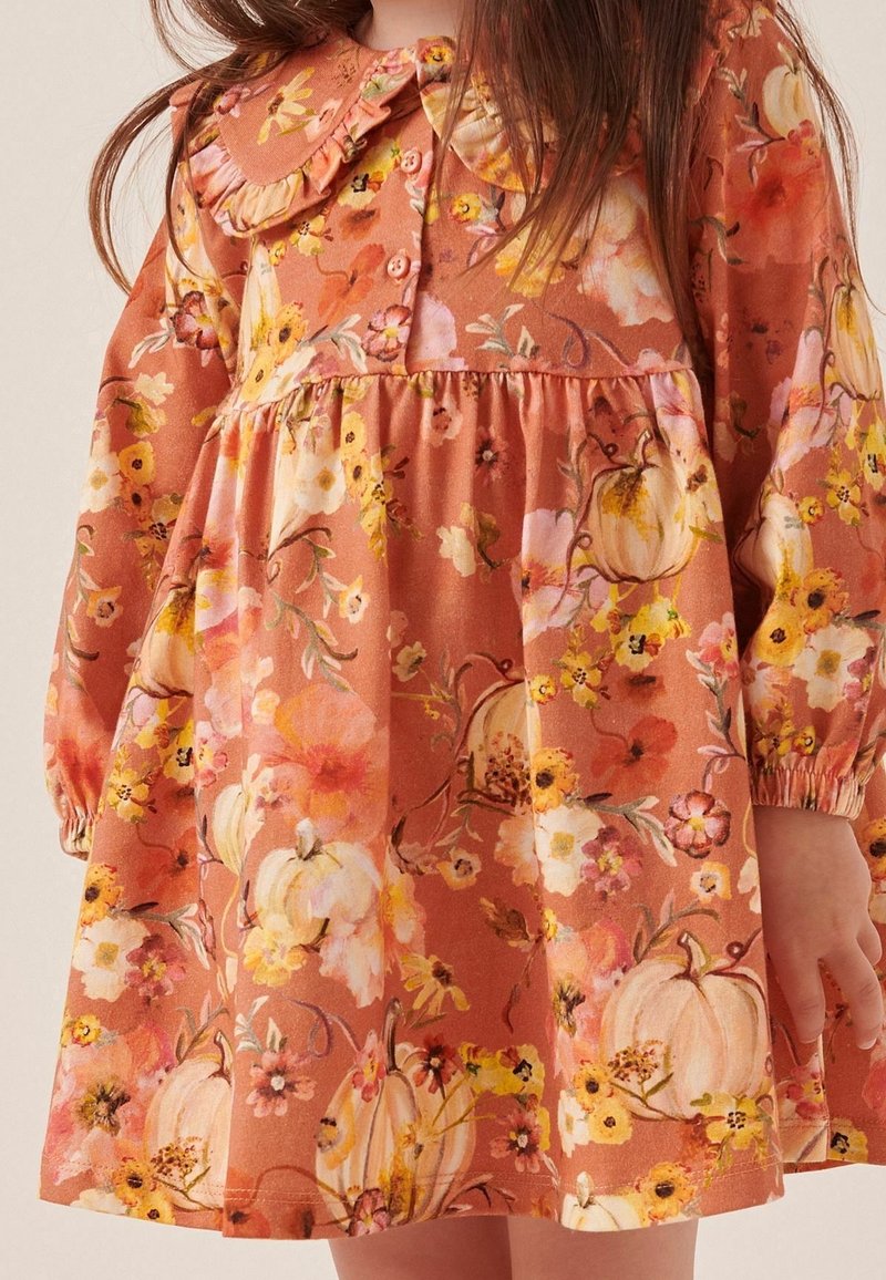 Enfant portant une robe orange rouille à manches longues, col à volants, avec un motif floral et de citrouilles blanches.