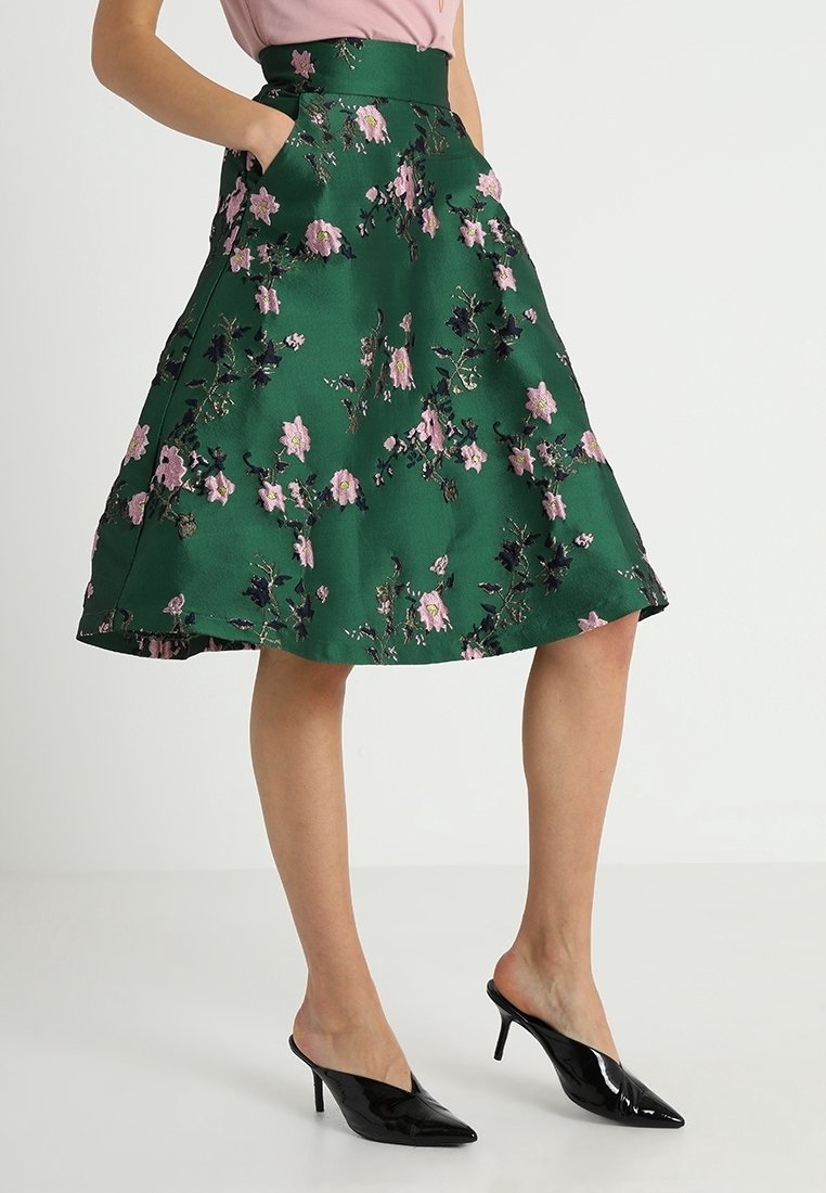 Jupe évasée verte avec broderie florale rose, taille haute, poches latérales, associée à des mules à talons noires à bout pointu.
