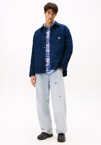 Jaunas vyras vilki tamsiai mėlyną siuvinėtą striukę ant mėlynos ruožinės marškinių, šviesius suplyšusius džinsus ir juodus sportbačius, stovintis prieš baltą foną.