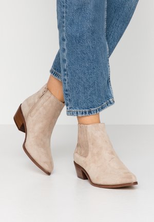 Hellbeige Wildleder-Stiefeletten mit seitlichem Reißverschluss und niedrigen hölzernen Blockabsätzen, getragen mit blauen Jeans.