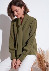 Olive green, long-sleeved blouse with a tied collar, gentle draping, and buttoned cuffs, paired with white pants, soft texture.