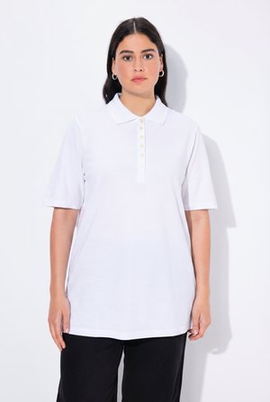 Frau mit dunklem Haar, die silberne Creolen-Ohrringe, ein weißes Poloshirt mit kurzen Ärmeln und Knöpfen sowie schwarze Hosen trägt, steht vor einem weißen Hintergrund.