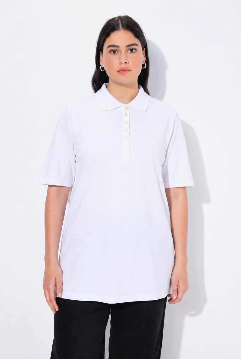 Frau mit dunklem Haar, die silberne Creolen-Ohrringe, ein weißes Poloshirt mit kurzen Ärmeln und Knöpfen sowie schwarze Hosen trägt, steht vor einem weißen Hintergrund.