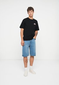 Black cotton t-shirt with short sleeves and a front graphic, paired with light blue denim shorts that reach the knee. White slip-on shoes.