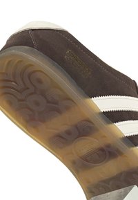 Καφέ σουέντ αθλητικό παπούτσι με τρεις λευκές ρίγες, υφή στη σόλα και λαστιχένια εξωτερική σόλα. Χρυσό λογότυπο "Gazelle" στο πλάι.