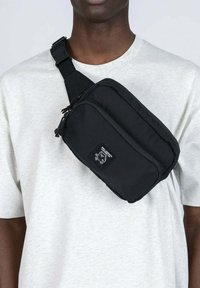 Henkilö, joka käyttää vaaleanharmaata t-paitaa, jossa on musta olkapääjauhe, jossa on etuvetoketjupussukka ja logopatch.