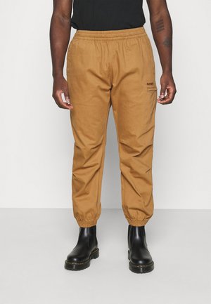 Homme portant un pantalon de jogging beige Levi's avec des poignets élastiques et des bottes en cuir noir, debout devant un fond uni.