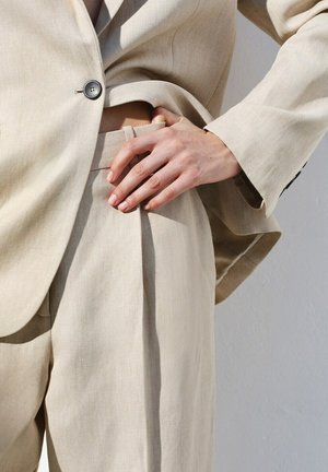 Primo piano di una persona che indossa un abito beige con una mano poggiata sulla vita, evidenziando la trama del tessuto e la vestibilità sartoriale.