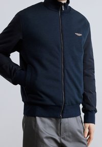 Personne portant une veste bleu marine zippée avec des poignets côtelés et un petit emblème ailé sur la poitrine gauche, associée à un pantalon gris.