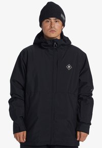 Svart vattentät jacka med dragkedja, huva och en diamantlogotyp på bröstet. Designen inkluderar justerbara ärmslut och fickor.