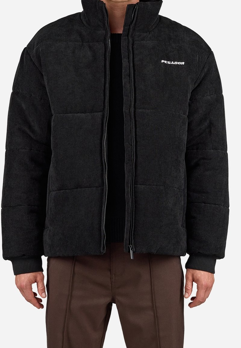 Veste matelassée noire en velours côtelé avec fermeture éclair à l'avant, col montant et logo blanc "PEGADOR" sur le haut gauche de la poitrine, portée par-dessus une chemise sombre et un pantalon marron.