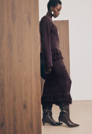 Mujer con un vestido texturizado de color púrpura oscuro con flecos y botas negras hasta la mitad de la pantorrilla, de pie de lado junto a paneles de madera.