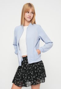 Veste bomber bleu clair à texture lisse, avec des poignets côtelés et une fermeture éclair. Associée à une jupe noire à motif floral.