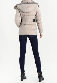 Mujer vestida con una chaqueta de plumas beige con capucha, jeans ajustados oscuros y botas negras hasta el tobillo, de pie de espaldas contra un fondo blanco.
