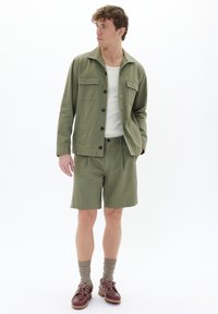 Jachetă verde măslinie cu două buzunare pe piept, combinată cu pantaloni scurți asortați; maiou alb dedesubt; pantofi maro cu șireturi de un bej deschis.