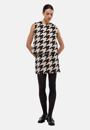 Un haut sans manches en motif pied-de-poule beige et noir, associé à une jupe assortie. Porté avec des collants noirs et des chaussures.