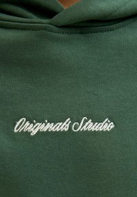 Tamnozelena dukserica s kapuljačom sa bijelim izvezenim natpisom "Originals Studio." Glatka tekstura s ležernim krojem i detaljima kapuljače.