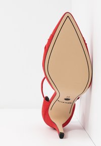 Chaussure à talon haut pointue rouge posée sur son talon, montrant une semelle beige lisse avec un contour noir et une indication de taille 37.