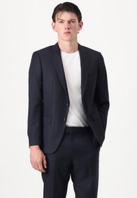 Jeune homme portant une veste de costume à fines rayures sombres et un pantalon assorti par-dessus un t-shirt blanc uni, se tenant devant un fond blanc.