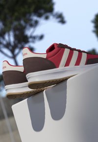 Paar rote und braune Adidas-Sneaker mit weißen Sohlen und Streifen, die auf einer weißen Oberfläche im Freien mit verschwommenen Bäumen im Hintergrund liegen.