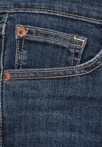 Mörkblå denimjeans med en framficka med gyllene sömnad och detaljer i kopparnitar. Texturen ser slät och robust ut.