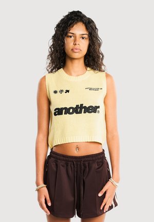 Młoda kobieta o kręconych włosach nosi żółtą bezrękawnikową dzianinową bluzkę z napisem „another” oraz czarne spodenki na jednolitym jasnym tle.