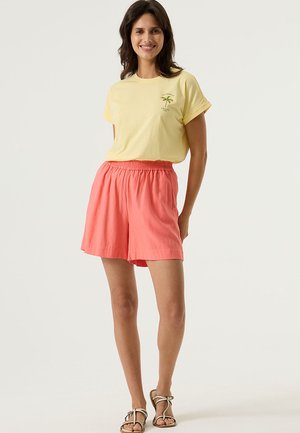 Lichtgeel katoenen T-shirt met een palmboomgrafiek, gecombineerd met losse koraalkleurige shorts met een elastische tailleband, gedragen met sandalen.