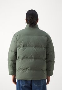 Person med mörkt hår i en knut, iklädd en olivgrön pufferjacka och blå jeans, visad från ryggen mot en enkel bakgrund.