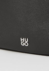 Svart teksturert skinnveske med sølvfarget "HUGO" logo på. Nærbilde viser strukturen i materialet og plasseringen av logoen.