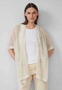 Gilet en crème léger et respirant à mailles ouvertes, avec des manches courtes, une coupe oversize et un bord côtelé. Porté par-dessus une chemise blanche ajustée.