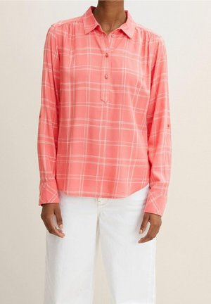 Femme portant une chemise à carreaux rose corail à manches longues avec des lignes blanches et un pantalon blanc, debout devant un fond uni.