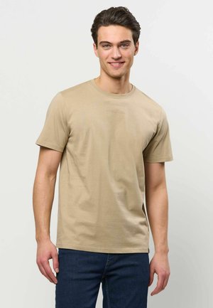 Junger Mann mit dunklem Haar, der ein beiges Kurzarm-T-Shirt und blaue Jeans trägt, lächelnd vor einem einfarbigen hellen Hintergrund.