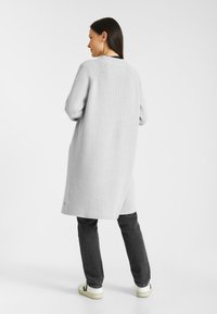 Lichtgrijze gebreide cardigan met een relaxte pasvorm, voorzien van een ronde halslijn, zijsplitten en ribbelstructuur, gedragen over donkere jeans en sneakers.
