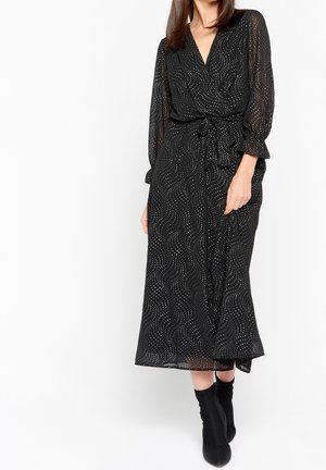 Femme portant une robe midi noire avec des motifs en spirale à pois blancs, des manches trois-quarts transparentes, une taille nouée, et des bottines noires.