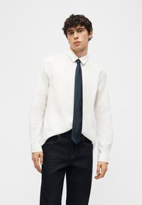 Νεαρός άνδρας με σγουρά μαλλιά, φοράει ένα λευκό πουκάμισο, σκοτεινή γραβάτα και μαύρο παντελόνι, στέκεται μπροστά σε απλό ανοιχτό φόντο.