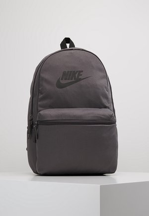Sac à dos Nike gris foncé avec poche avant zippée et logo noir, posé sur un socle blanc sur fond gris clair.