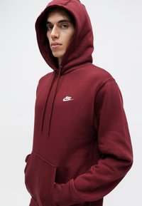Bordová mikina s kapucňou a predným vreckom, s bielym logom Nike na hrudi. Vyrobená z mäkkého, textúrovaného materiálu.
