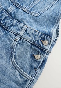 Lichtblauwe denim overall met een licht verweerde textuur, twee metalen knopen en zijzakken met zichtbare stikdetails.