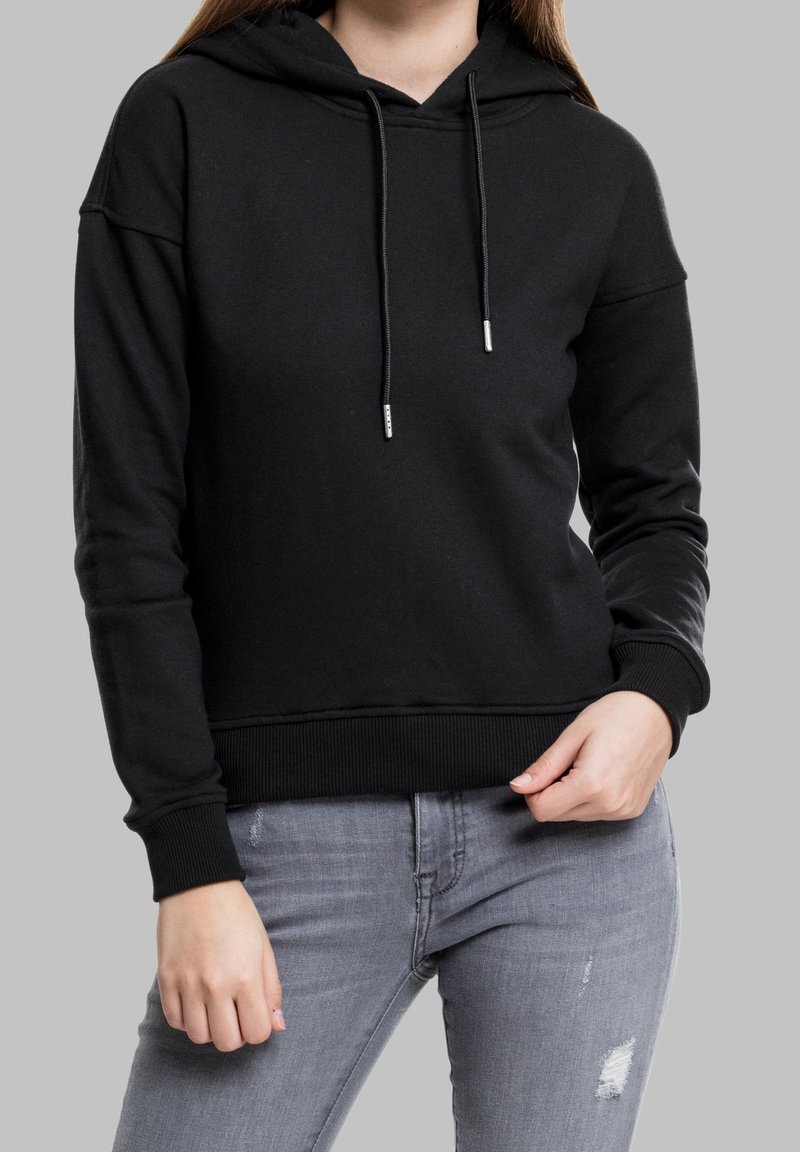 Femme portant un sweat à capuche noir uni avec cordons et un jean gris clair déchiré, debout devant un fond gris.
