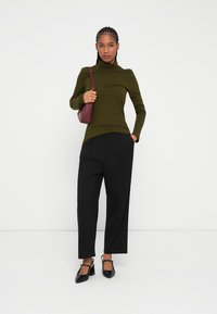 Pull à col roulé vert, pantalon noir à large jambes, chaussures noires. Petit sac à main bordeaux. Le mannequin se tient devant un fond blanc uni.