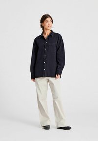 Dunkelblaue Denimhemd mit Knopfleiste, spitzem Kragen und einer einzigen Brusttasche, kombiniert mit locker sitzenden beigen Hosen und schwarzen Schuhen.