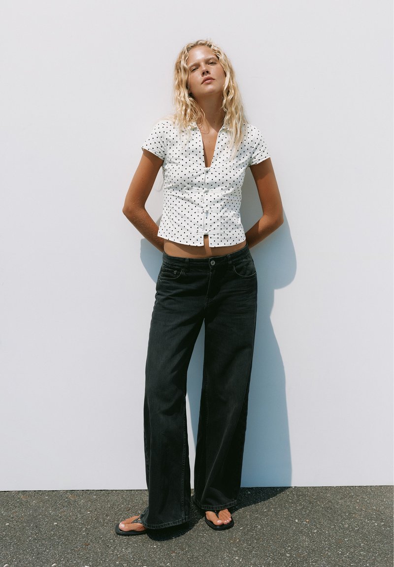 Jeune femme aux longs cheveux blonds portant une chemise blanche à manches courtes à pois, un jean noir à jambes larges et des tongs noires, debout contre un mur blanc.