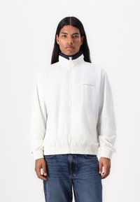White zip-up jacket with a high collar and elastic cuffs. Features a navy collar detail and subtle branding on the chest. Worn over blue jeans.