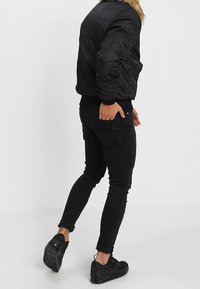 Černá péřová bunda s kožešinovým límcem, v kombinaci s černými skinny džíny se strukturovanými kapsami a černými teniskami. Boční pohled na outfit.