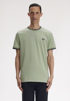 Homme portant un t-shirt à manches courtes vert clair avec bordure noire et logo de laurier, debout devant un fond blanc uni.