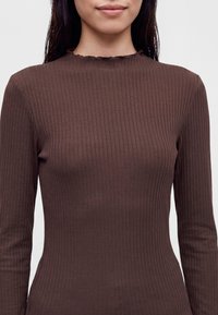 Femme portant un haut marron moulant en maille côtelée à manches longues avec un col montant subtilement volanté sur un fond uni.