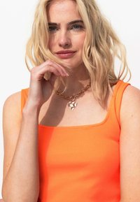 Débardeur orange à rayures avec un col carré, associ é à un collier en chaîne dorée avec un pendentif en forme de cœur. Fond clair.