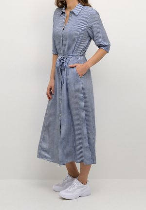 Woman wearing a blue and white vertical striped midi shirt dress with half sleeves, a tied waist belt, and white sneakers.