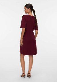 Abito a maglia bordeaux con maniche corte e un fit rilassato, che termina sopra il ginocchio. Indossato con sandali con tacco marroni. Texture liscia, design minimal.