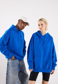 To personer har på seg oversized blå hettegensere laget av myk tekstil, med kengurulommer og justerbare snorer.